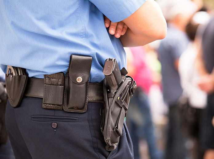 A police officer in uniform stands with arms crossed, showing a utility belt with a holstered gun and other equipment; blurred people are visible in the background.