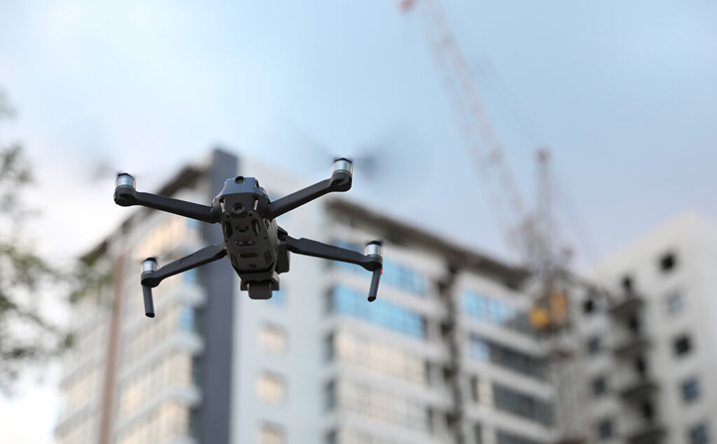 A black drone flying outdoors in front of a modern multi-story building, with a construction crane visible in the background under a partly cloudy sky.