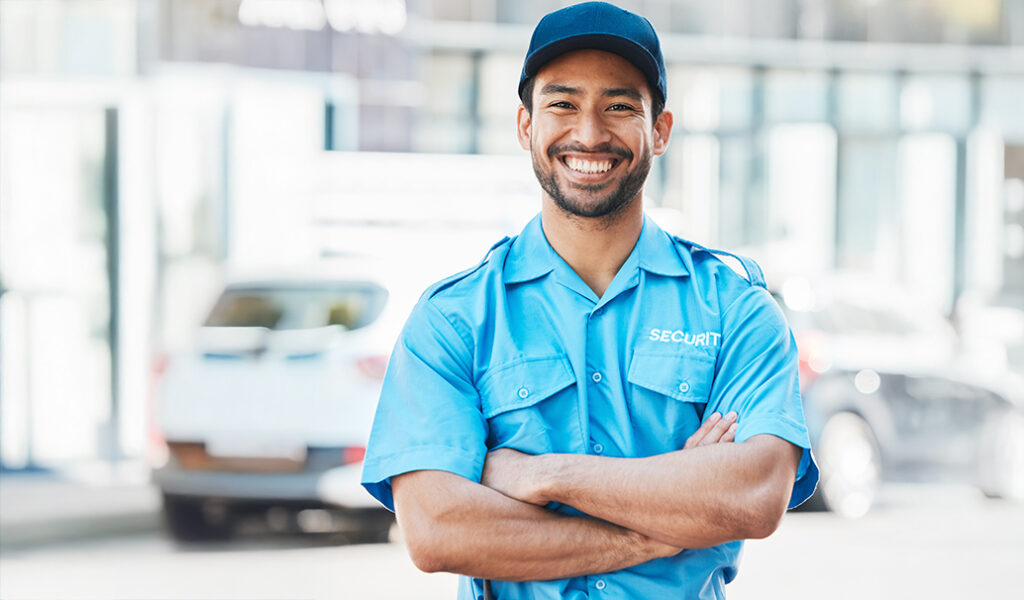 A smiling security guard in a blue uniform and cap stands outdoors with arms crossed, in front of blurred vehicles and buildings.