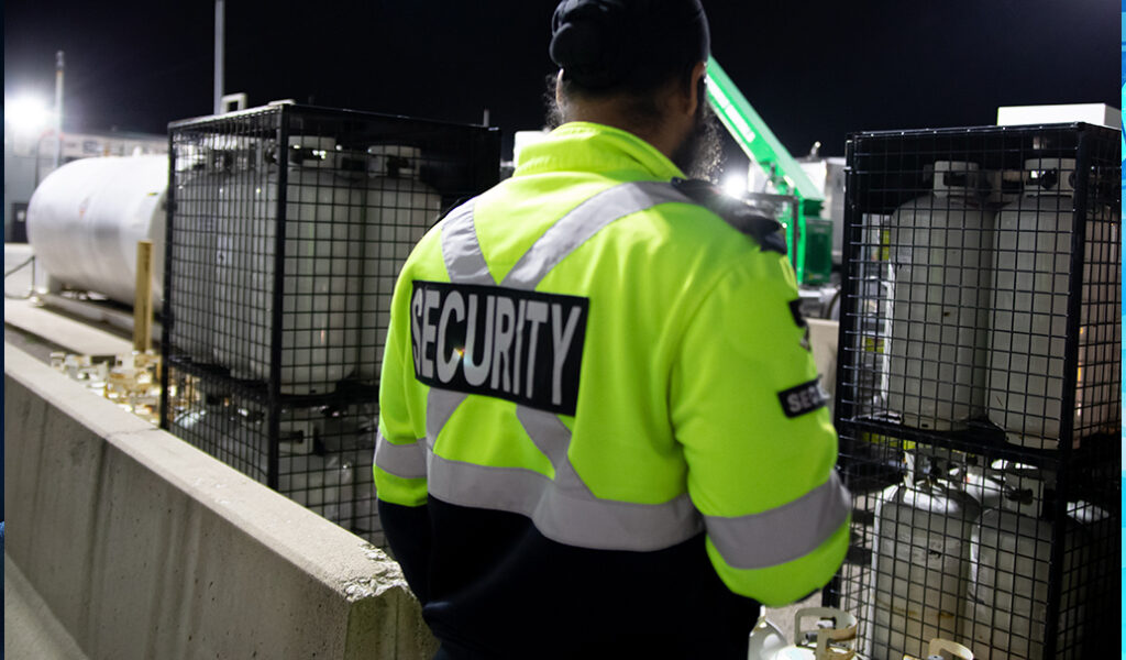 A security guard wearing a high-visibility jacket stands near large storage tanks at night, monitoring the area.