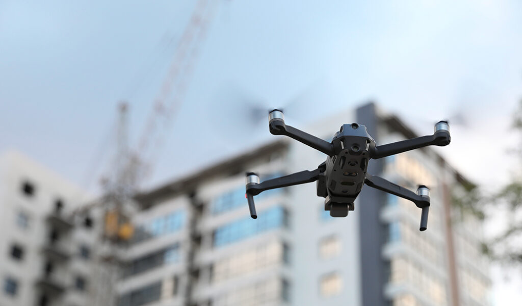 A black drone hovers in the foreground with a blurred modern apartment building and a construction crane visible in the background.
