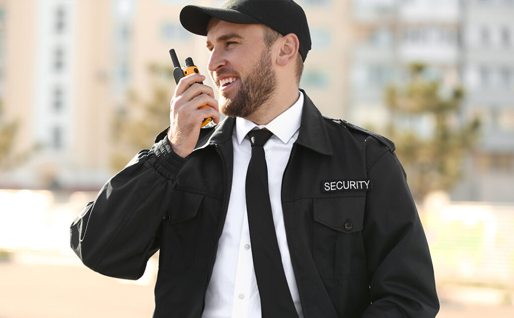 A male security guard in a black uniform and cap holds a walkie-talkie to his mouth, smiling outdoors with blurred buildings in the background.