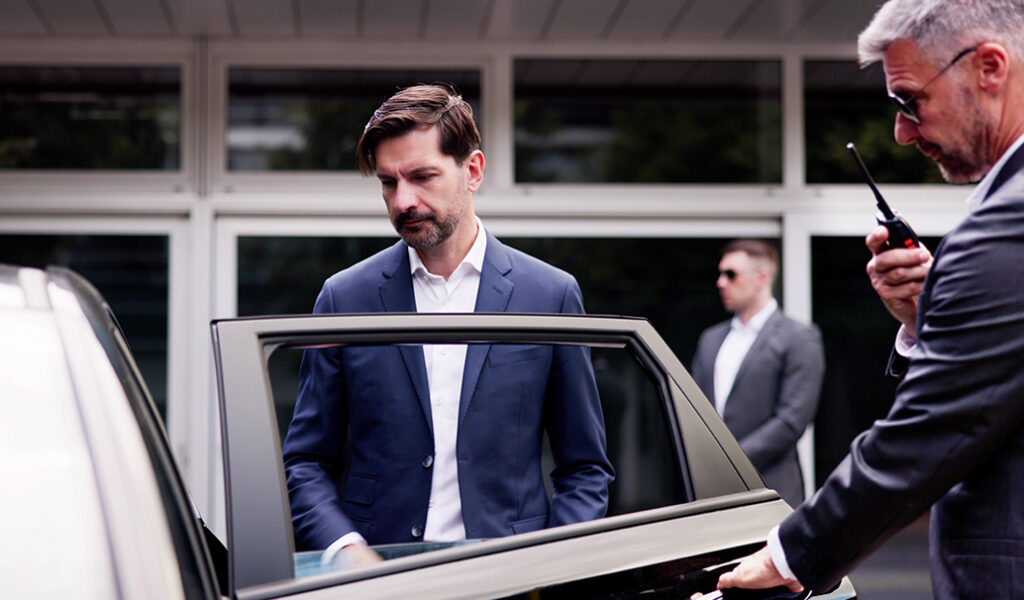 A man in a suit prepares to enter a car while another man, also in a suit and sunglasses, holds the car door open. Another person in sunglasses stands in the background, suggesting a security detail.