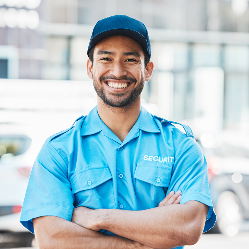 A smiling security guard wearing a blue uniform and cap stands with arms crossed in an outdoor setting, with blurred buildings and cars in the background.