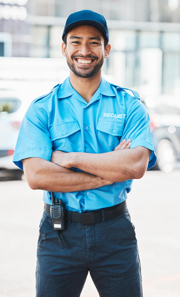 A smiling security guard in a blue uniform and cap stands outdoors with arms crossed, a walkie-talkie attached to his belt, and blurred cars and buildings in the background.
